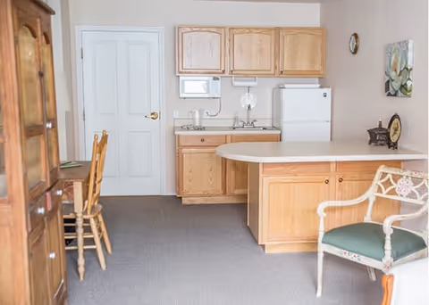 A small kitchen area with light wood cabinets, a white refrigerator, a microwave, and a sink. There is a countertop extending into the room with a decorative chair nearby. To the left, there is a wooden desk with two wooden chairs. The walls are light-colored, and there is a clock and a framed picture hanging on the wall.