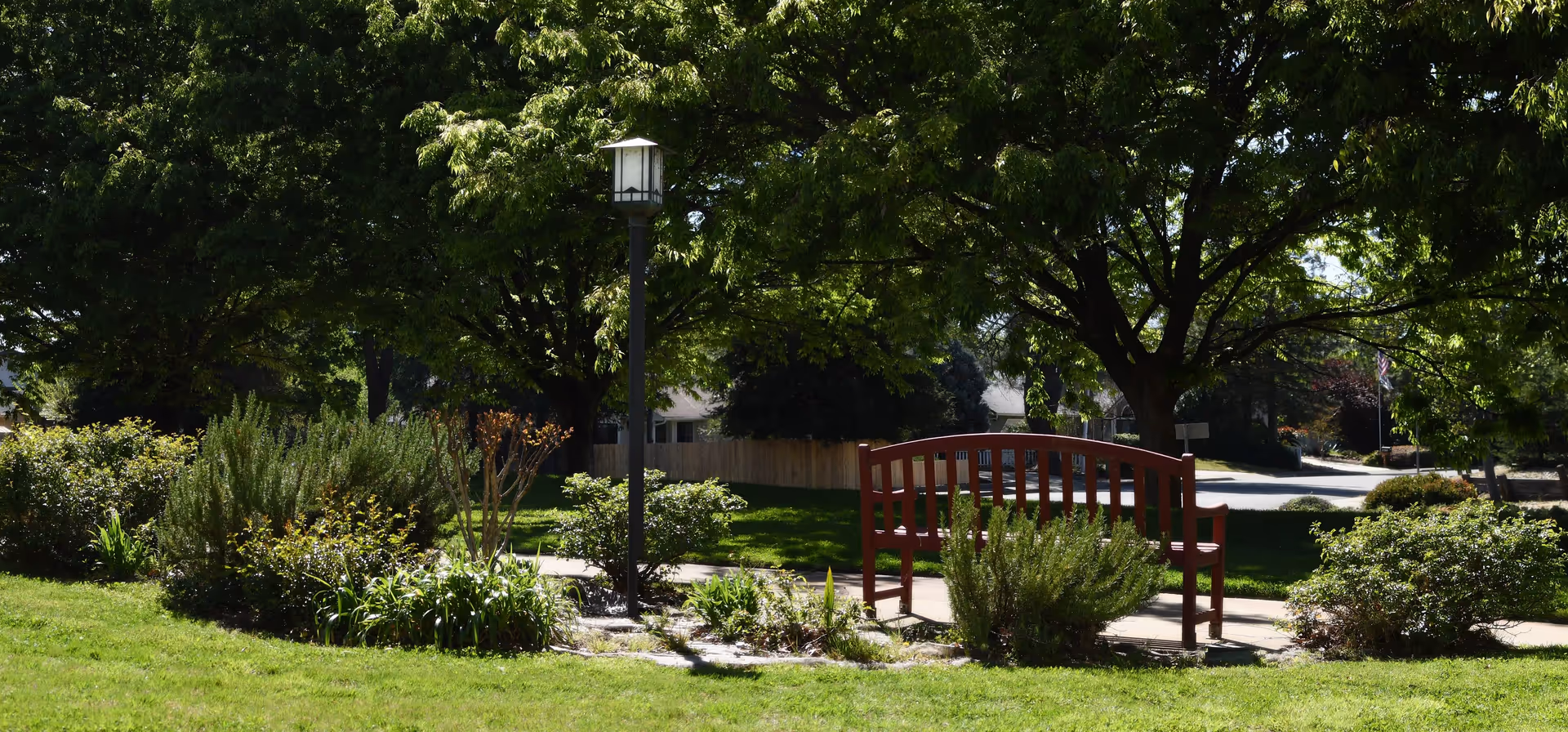 A peaceful outdoor garden area with a wooden bench, surrounded by green grass, bushes, and large leafy trees providing shade. A black lamp post stands near the bench along a paved pathway.