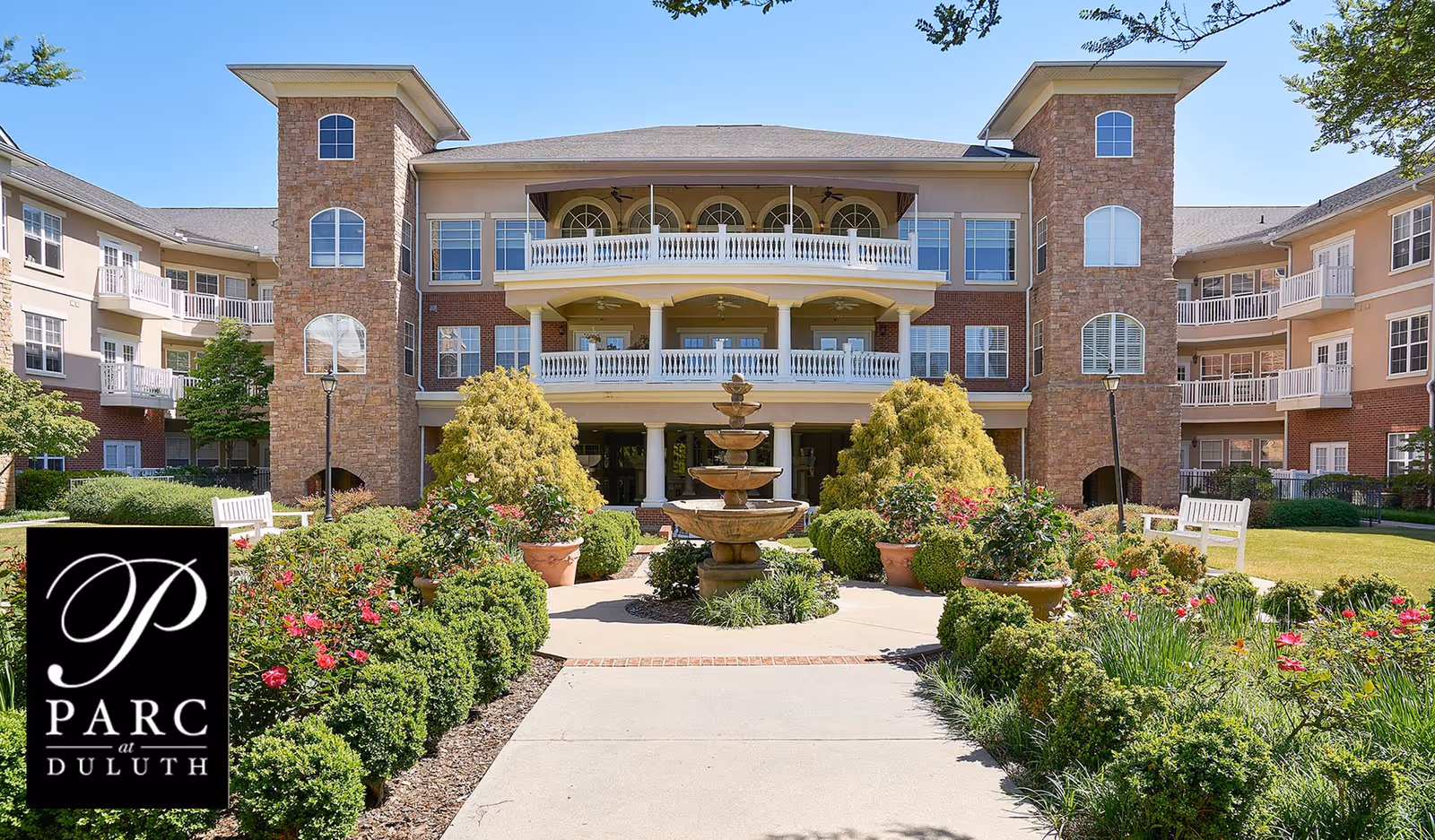 Landscaped courtyard with a central fountain in front of a three-story senior living building with balconies.