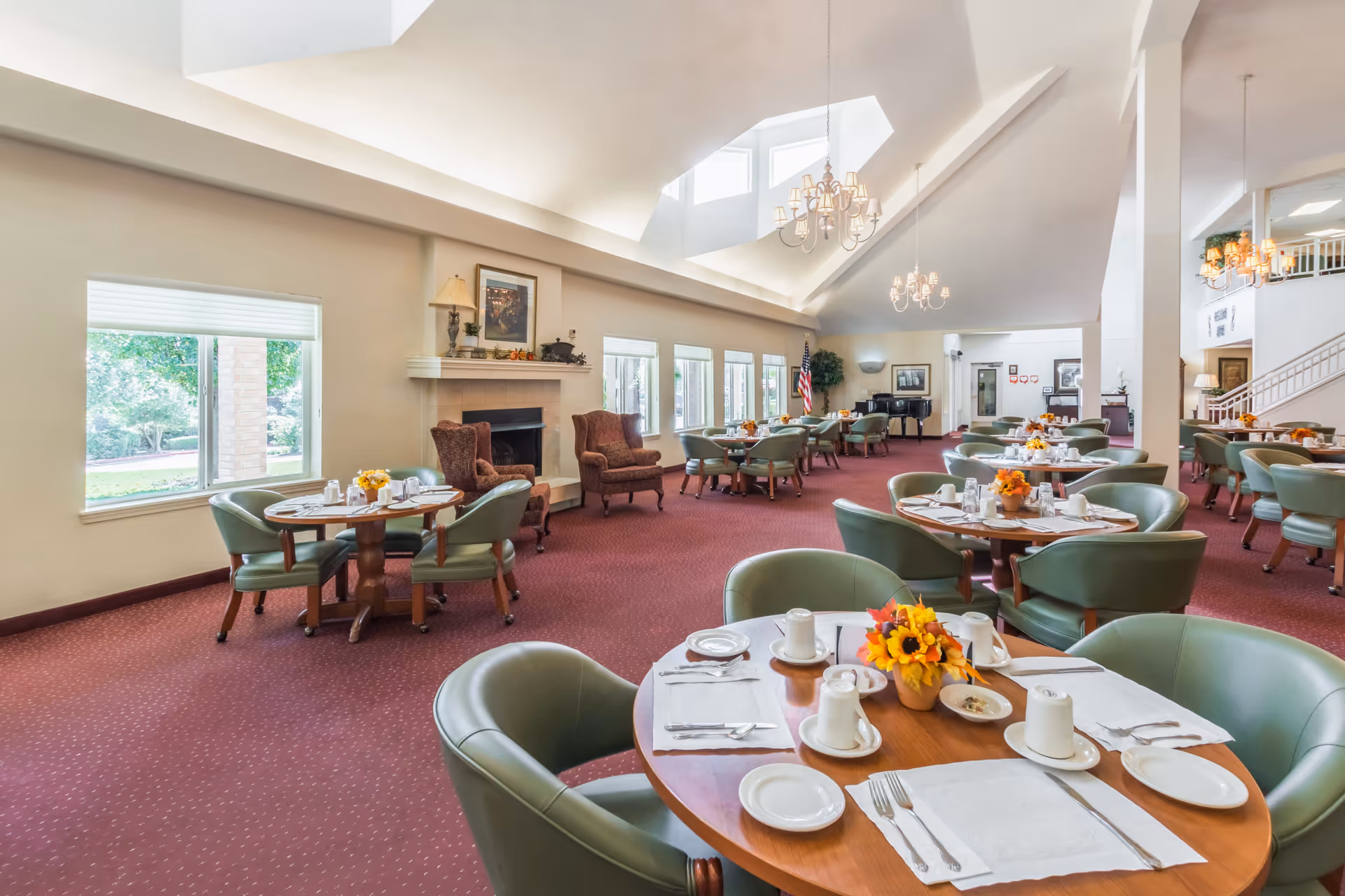 A spacious dining room with multiple round wooden tables set with white plates, cups, and silverware. Each table has a small floral centerpiece with yellow and orange flowers. The room has green cushioned chairs around the tables, a red carpeted floor, large windows letting in natural light, a fireplace with two brown armchairs, chandeliers hanging from a high ceiling, and an American flag near a black grand piano in the background.