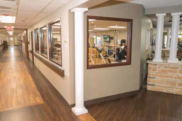 Interior hallway with wood-look floors, decorative columns and windows into a therapy/activity room.