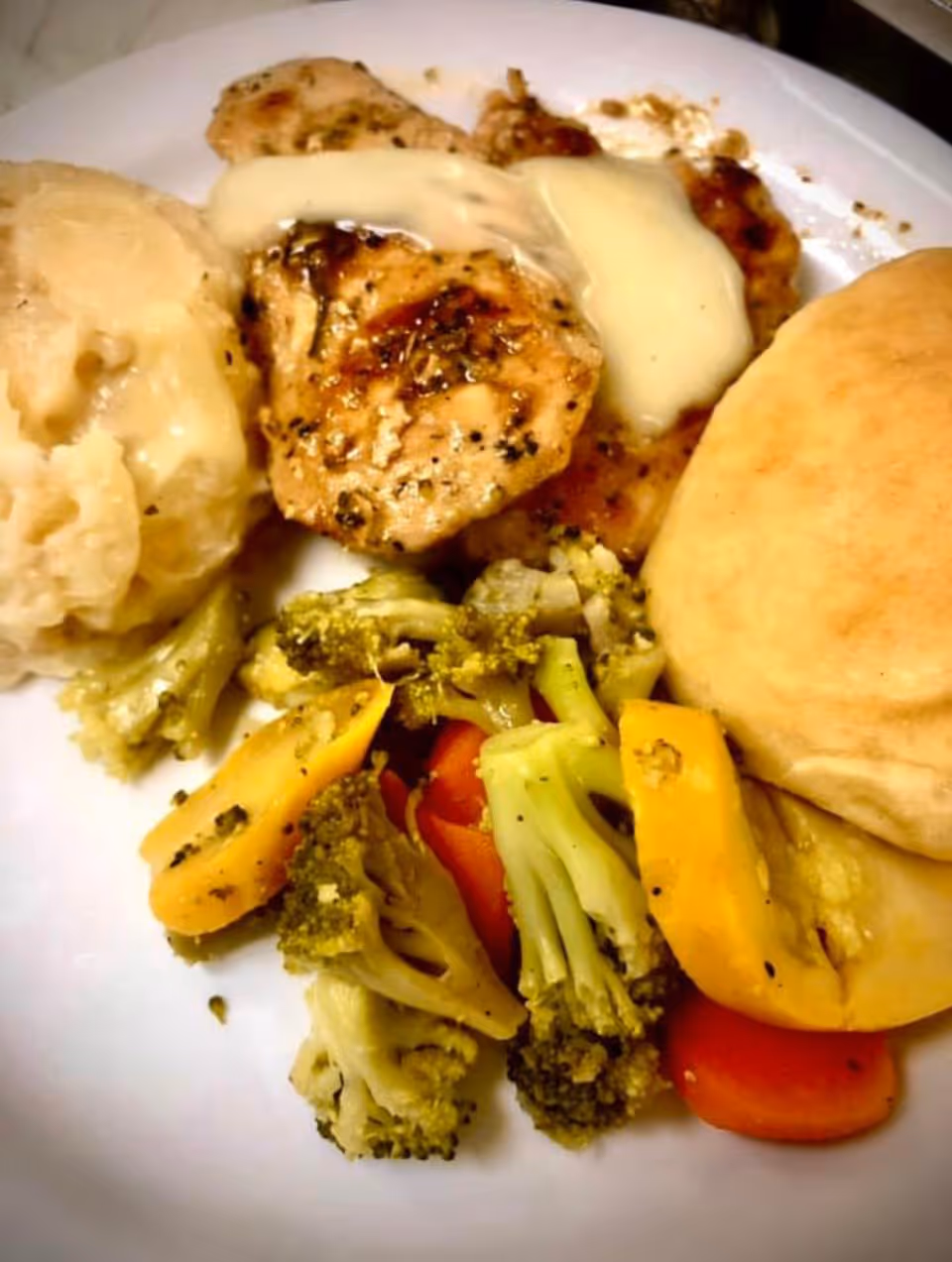 A plate of food featuring grilled chicken breast topped with a creamy sauce, mashed potatoes with gravy, steamed mixed vegetables including broccoli, carrots, and yellow squash, and a bread roll.
