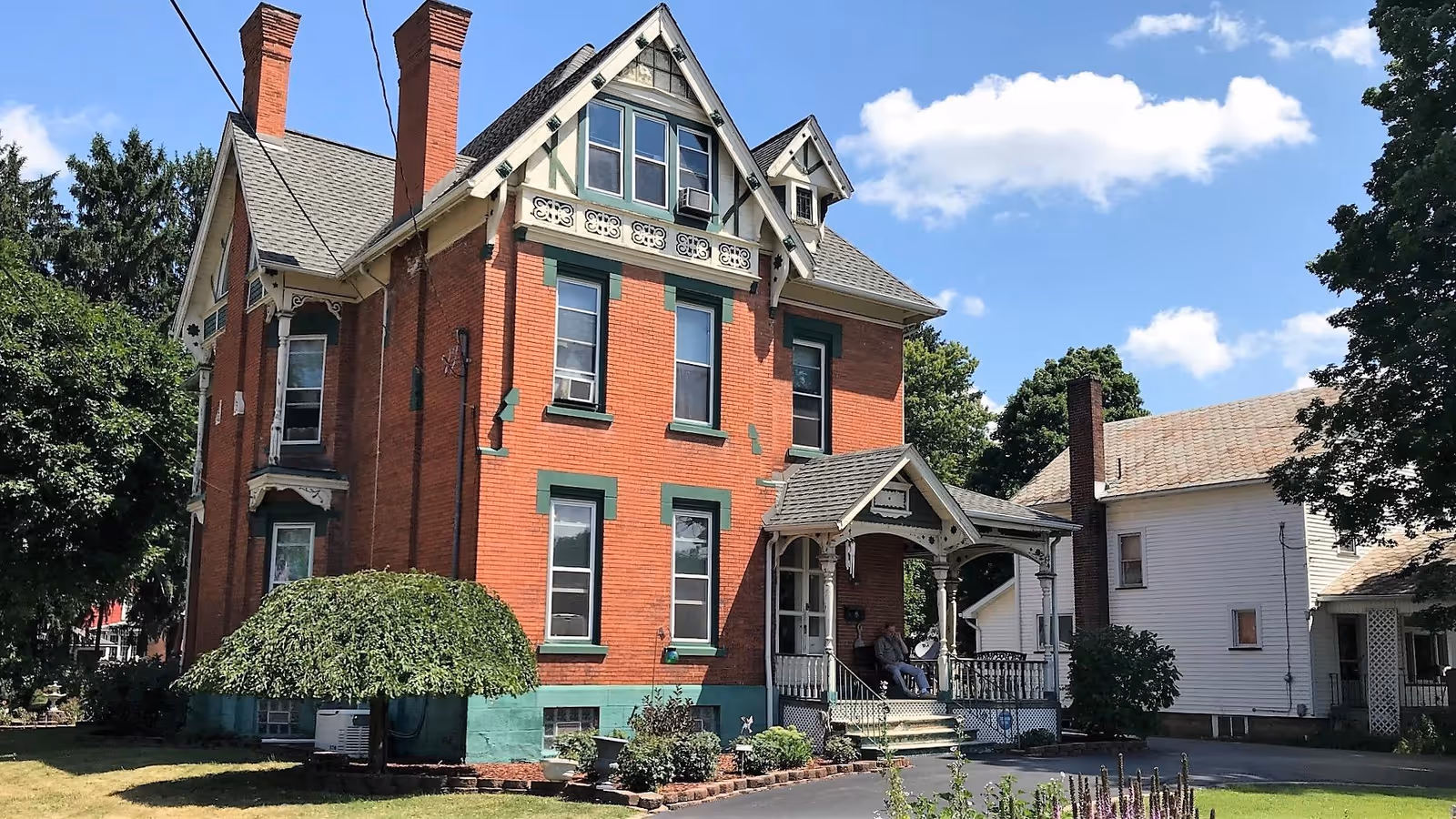 Front exterior of a red-brick Victorian house with green trim and a covered porch under a blue sky.