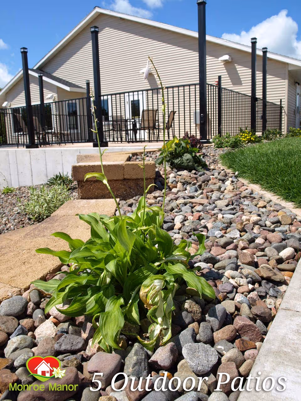 Outdoor patio area at Monroe Manor featuring a rock garden with green plants in the foreground, a concrete walkway, and a fenced patio with tables and chairs attached to a beige building under a blue sky with some clouds.