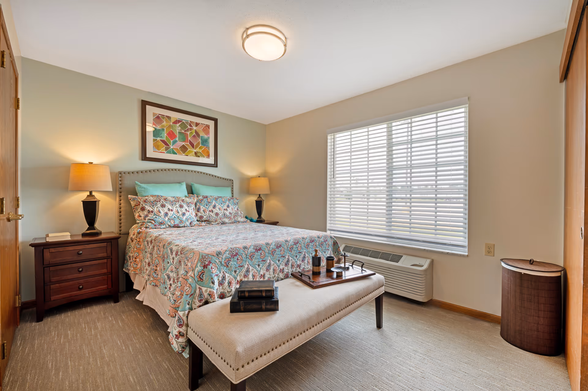 Well-lit bedroom with a patterned bed, two bedside tables with lamps, a bench at the foot of the bed, and a large window with blinds.