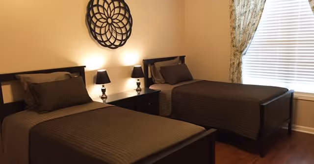 A bedroom with two single beds featuring dark wooden frames and matching brown bedding. Between the beds is a nightstand with two black lamps. Above the nightstand is a decorative circular wall art. A window with blinds and patterned curtains is on the right side of the room.
