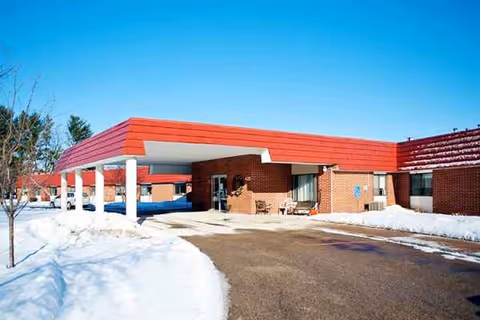 Snow surrounds a red-roofed senior living facility entrance with brick walls and a clear blue sky.
