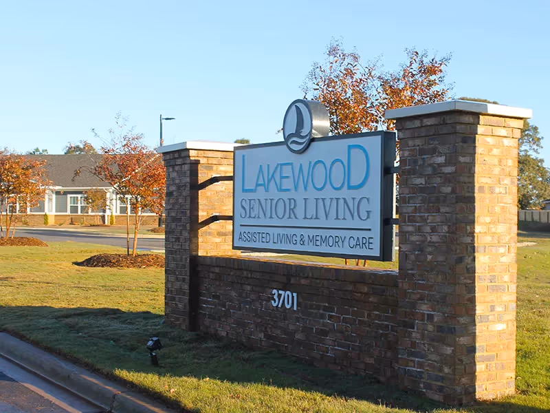 A brick sign at the entrance of Lakewood Senior Living facility, displaying the name and services offered including assisted living and memory care, with a building and trees in the background.