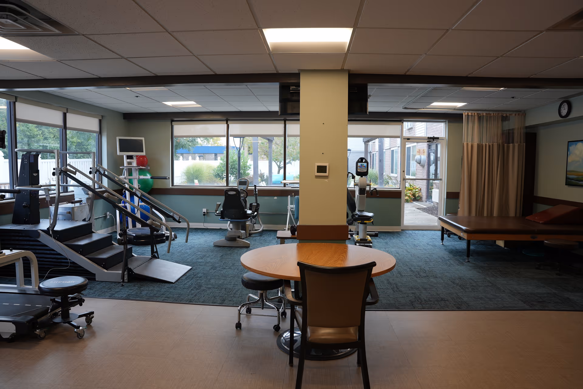 A rehabilitation or physical therapy room with exercise equipment including a treadmill, stair stepper, and stationary bikes. There is a round wooden table with chairs in the foreground, large windows letting in natural light, and a therapy table with a curtain divider on the right side. The room has a carpeted area and a tiled floor section.