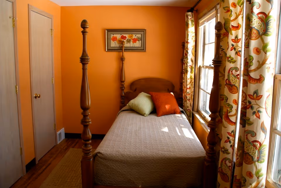 A cozy bedroom with orange walls featuring a wooden four-poster bed with a beige quilt and two decorative pillows, one green and one orange. There is a framed floral artwork on the wall above the bed, and a window with patterned curtains allowing natural light to enter the room. Two closed wooden doors are visible on the left side.