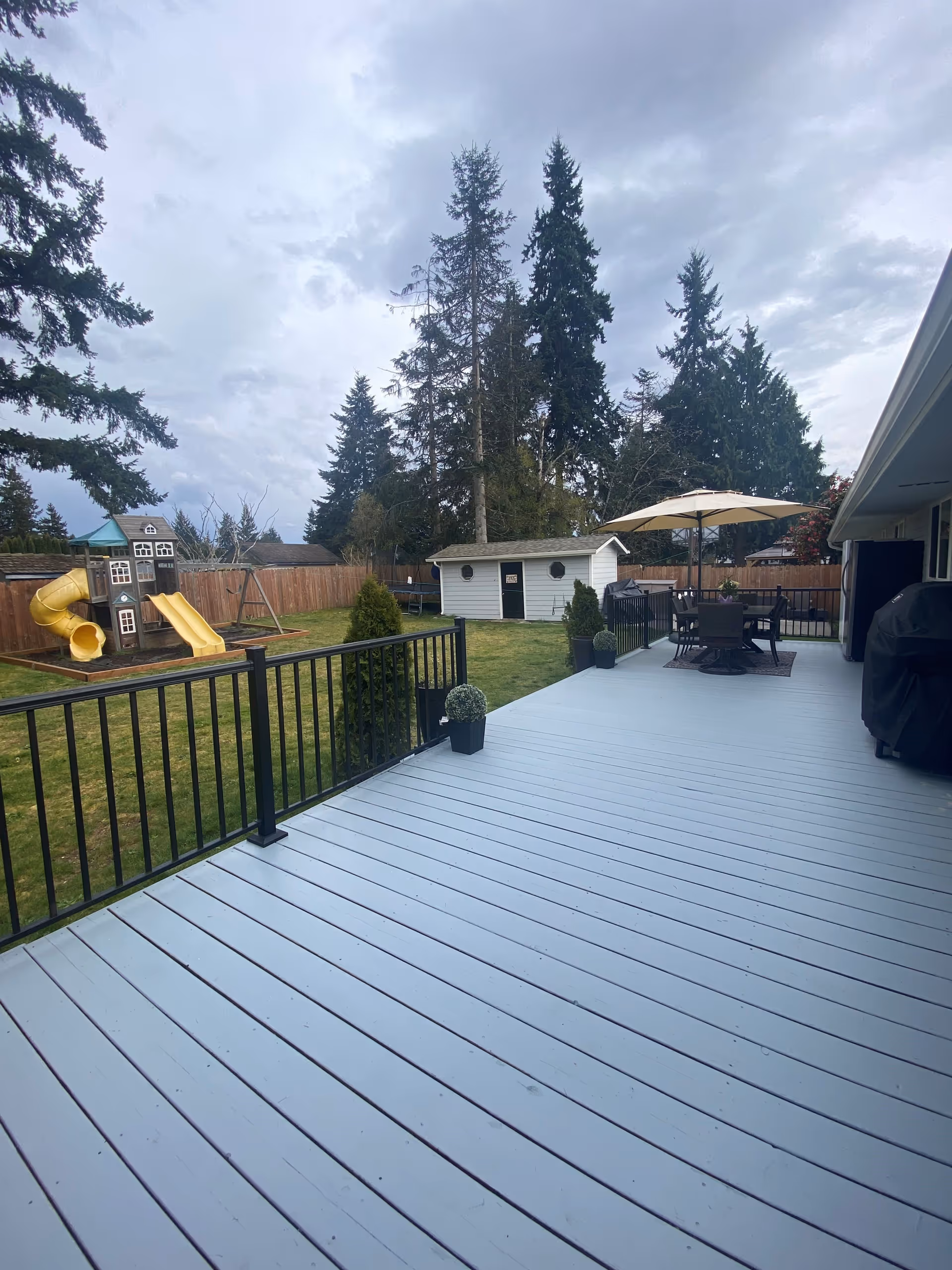 A spacious outdoor deck area with light blue wooden flooring and black metal railing. The deck has a round table with chairs and a large umbrella. Beyond the deck is a grassy yard with a children's play structure featuring slides and swings, a small white shed, and tall evergreen trees in the background under a cloudy sky.