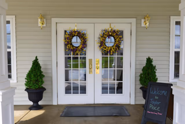 Double white glass-paneled doors decorated with two wreaths made of purple and yellow flowers. On either side of the doors are black urn planters with green topiary plants. A blackboard sign on the right side reads 'Welcome to Taylor Place'. The exterior wall is light gray with white trim and two gold wall-mounted lanterns above the planters.