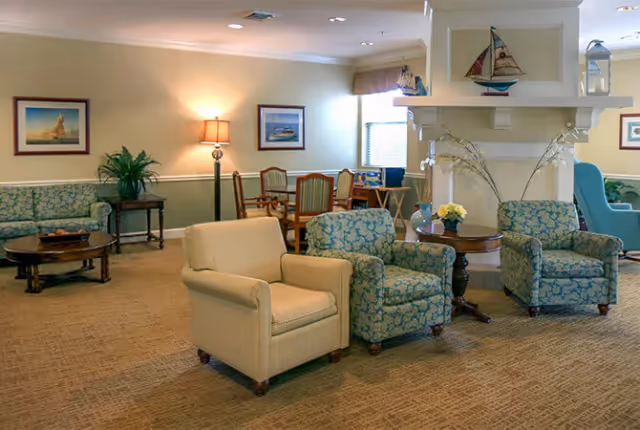 A cozy living room area with beige and patterned armchairs arranged around wooden tables. The room features soft lighting from a floor lamp, framed nautical-themed artwork on the walls, and decorative sailboat models on a white mantel. In the background, there is a dining area with wooden chairs and a window letting in natural light.