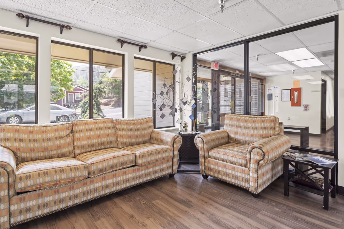 Sunlit seating area with a patterned sofa and armchair in a facility lobby with large windows and wood flooring.