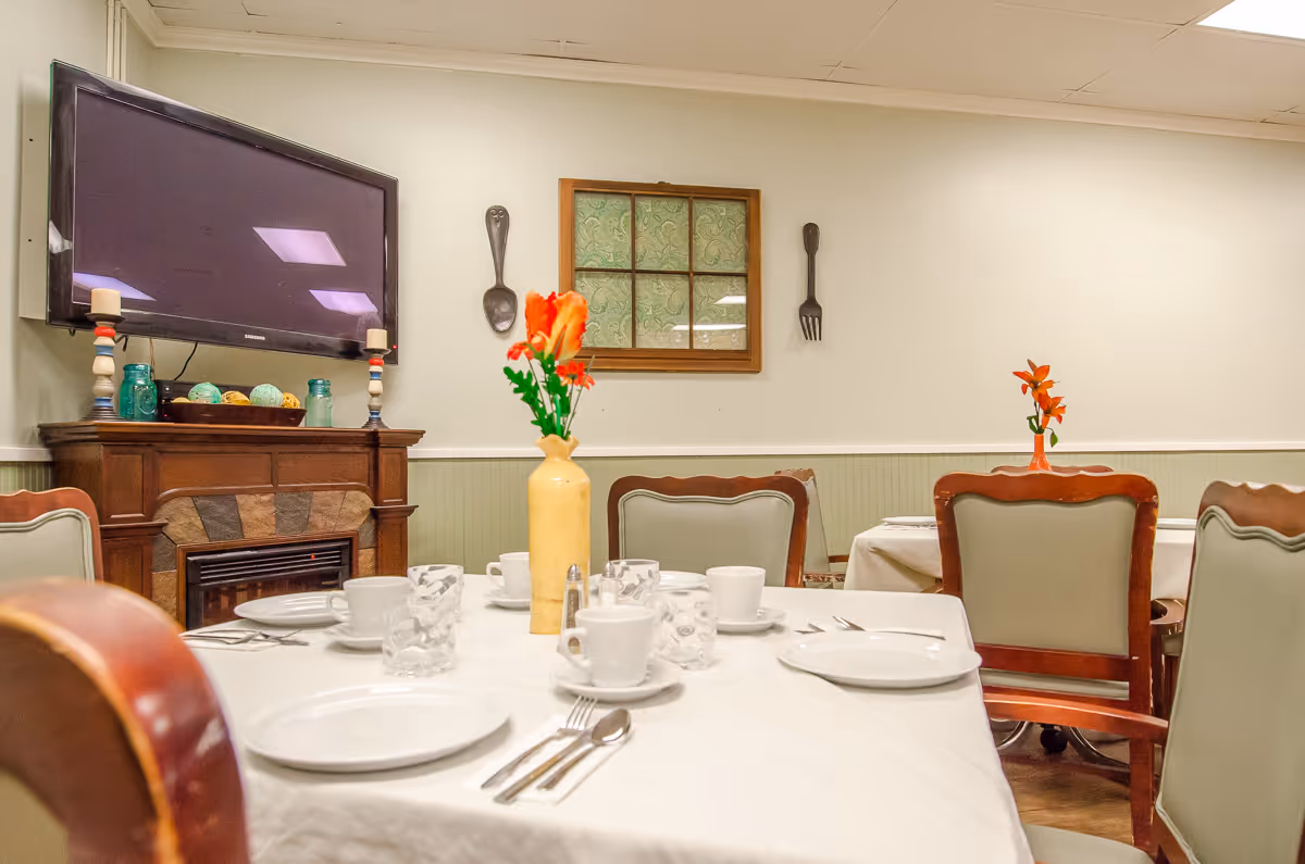A small dining room set for a meal with white-tableclothed tables, place settings, vases of flowers, chairs, and a wall-mounted TV.