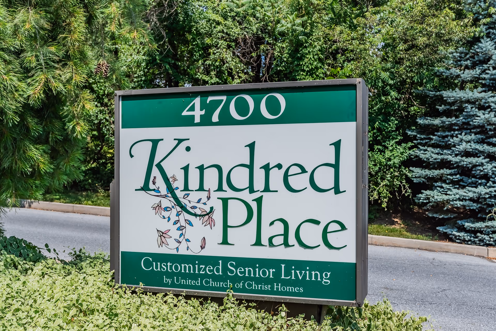 A green and white outdoor sign for Kindred Place, a customized senior living facility by United Church of Christ Homes, located at 4700. The sign is surrounded by greenery and trees.