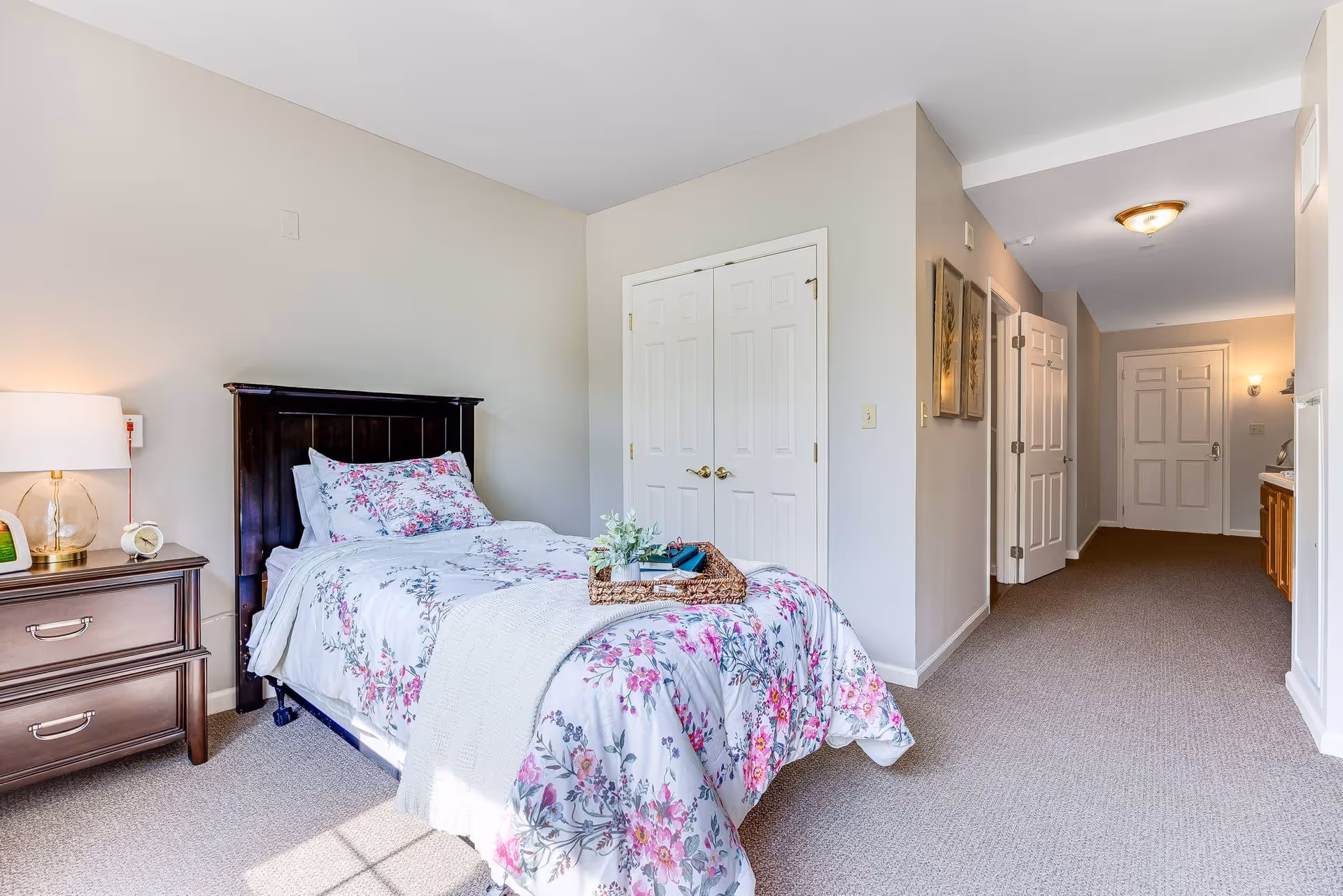 Bright senior living bedroom with a single bed dressed in floral bedding, a nightstand, double closet doors, and a hallway leading to the entrance.