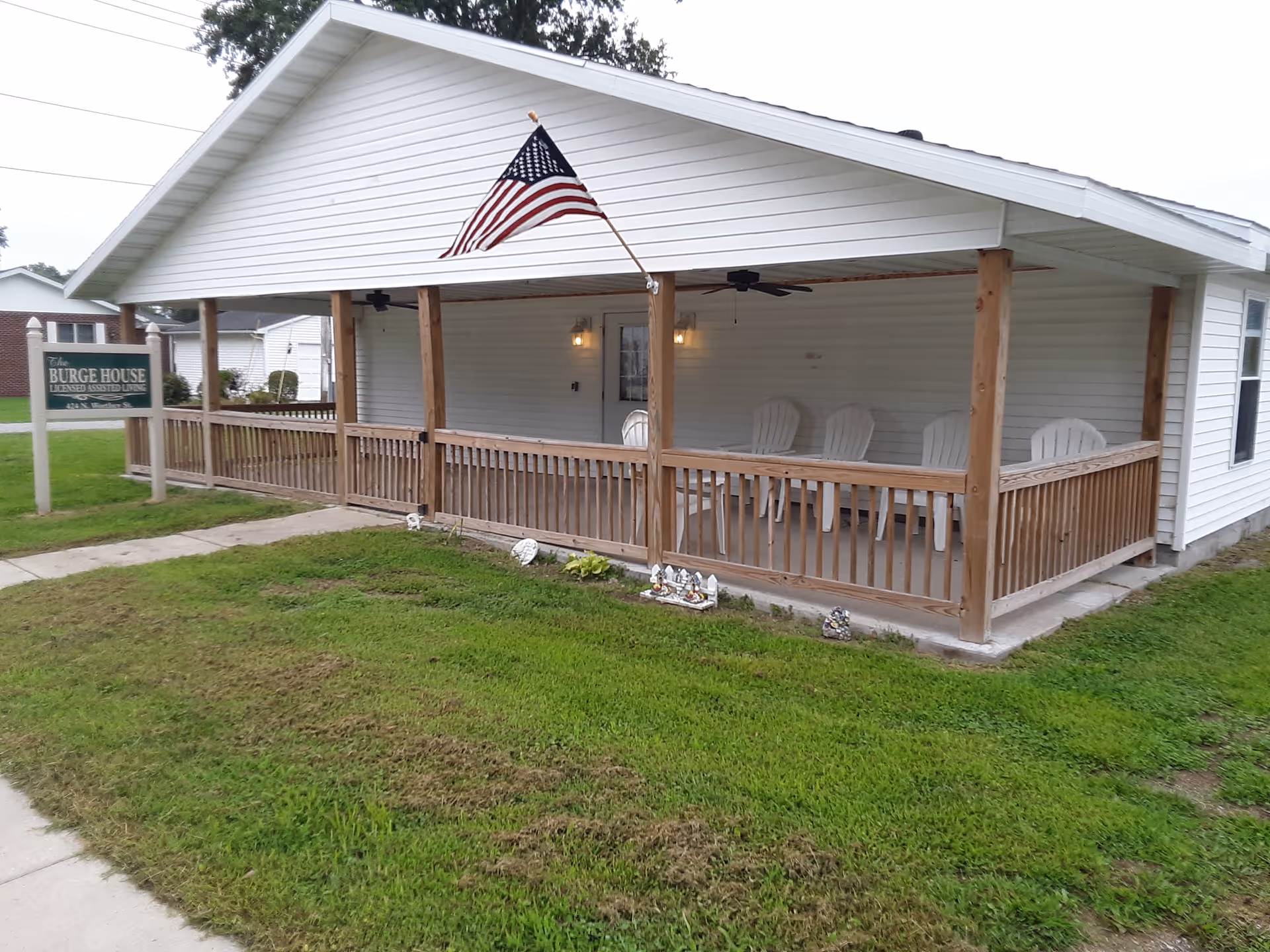 Front view of a single-story white building with a covered wooden porch, several chairs, an American flag, and a sign reading 'Burge House'.