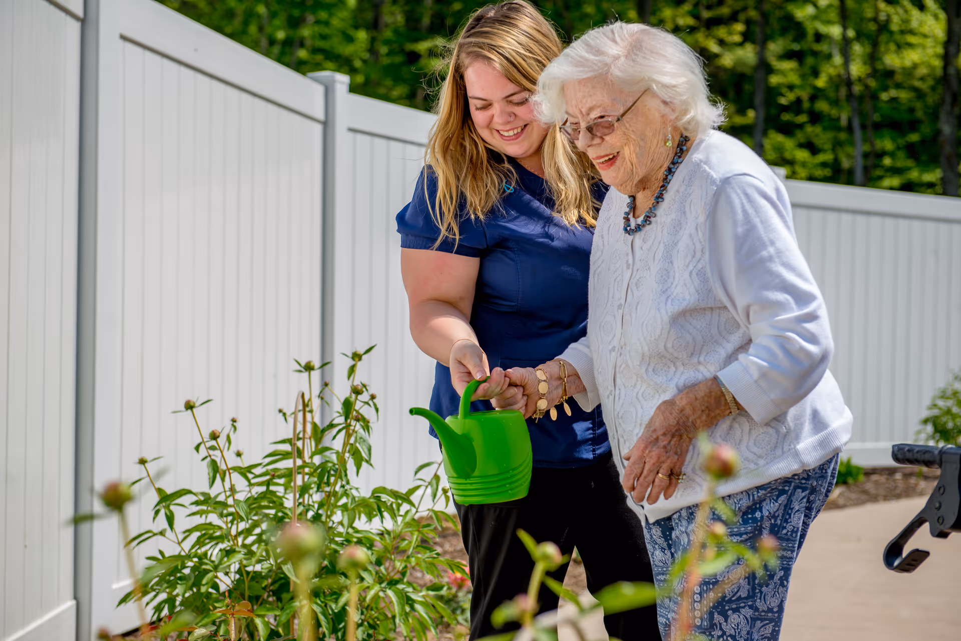 A young female caregiver helps an elderly woman water plants in a garden outside, both smiling and enjoying the activity near a white fence.