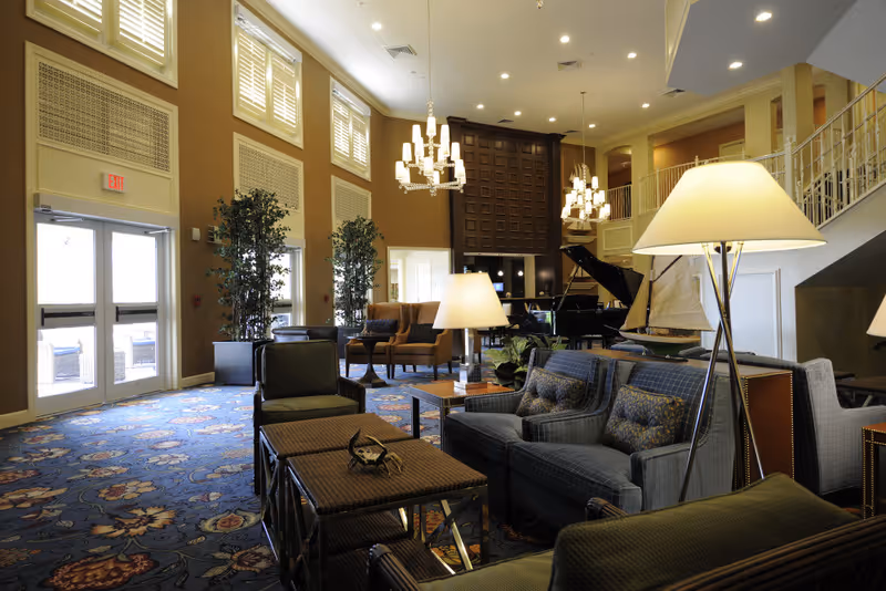 A spacious and well-lit living room area with multiple seating options including sofas and armchairs arranged around coffee tables. The room features tall windows with shutters, decorative chandeliers, potted plants, a grand piano, and a staircase leading to an upper level. The carpet has a floral pattern and there are several floor lamps providing warm lighting.
