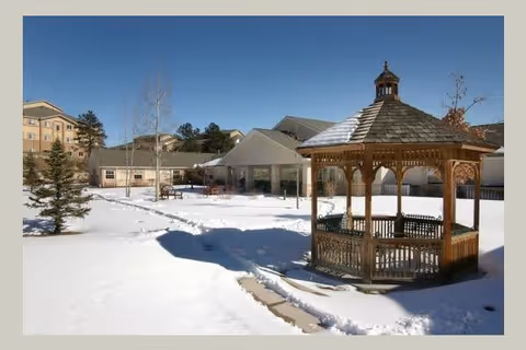 Snow-covered outdoor area of a senior living facility with a wooden gazebo in the foreground and buildings in the background under a clear blue sky.