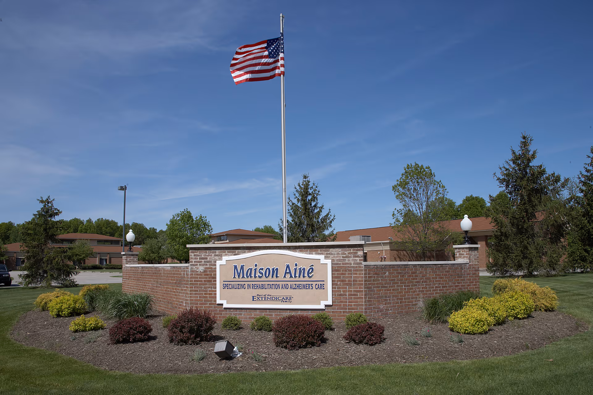 Outdoor view of a brick sign for Maison Ainé, specializing in rehabilitation and Alzheimer's care, with an American flag flying on a pole behind it. The sign is surrounded by landscaped bushes and grass, with trees and buildings in the background under a clear blue sky.