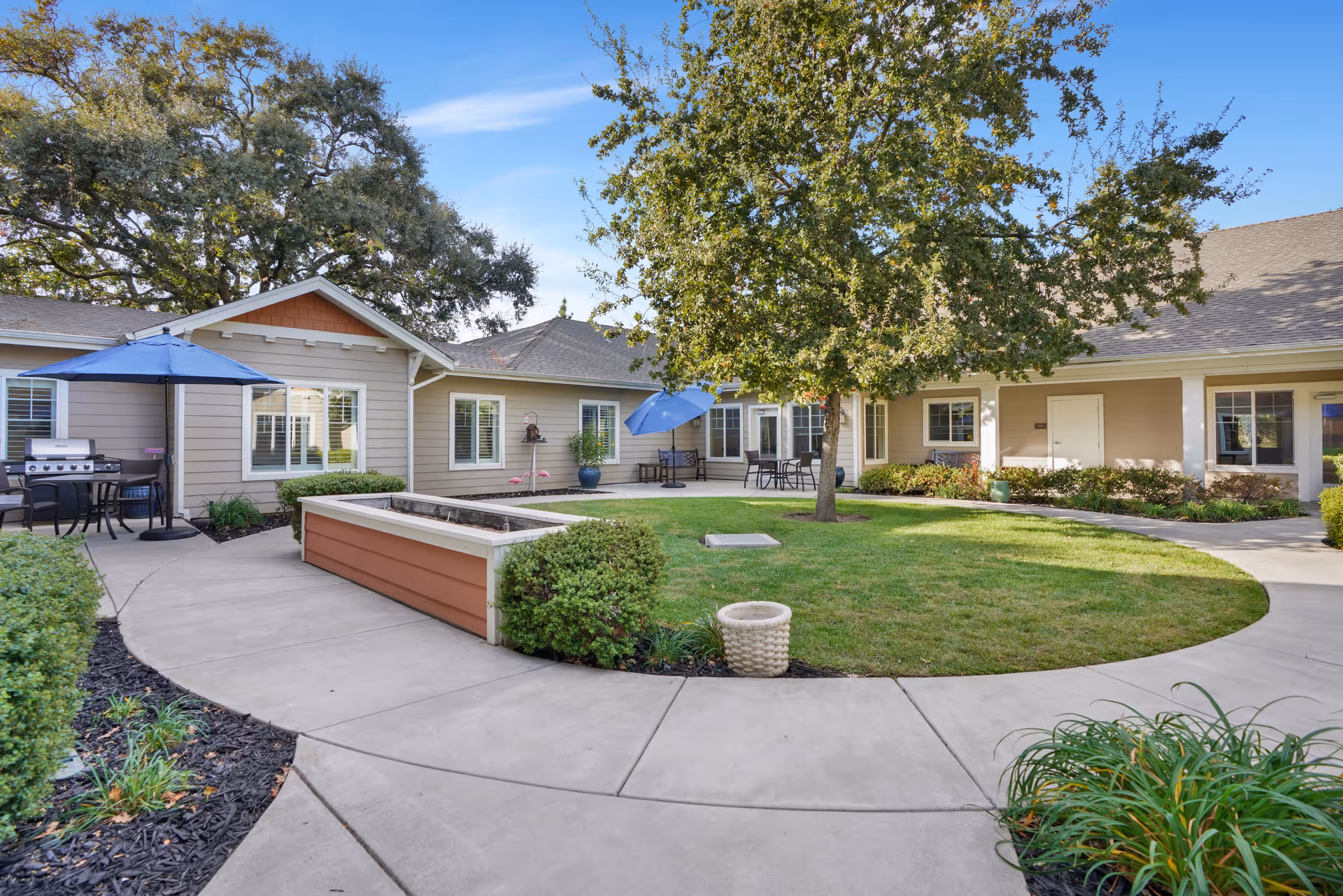 Outdoor courtyard area at The Commons At Elk Grove featuring a circular grassy lawn with a tree in the center, surrounded by a concrete walkway. There are patio tables with blue umbrellas, a barbecue grill, and beige buildings with white trim in the background under a clear blue sky.