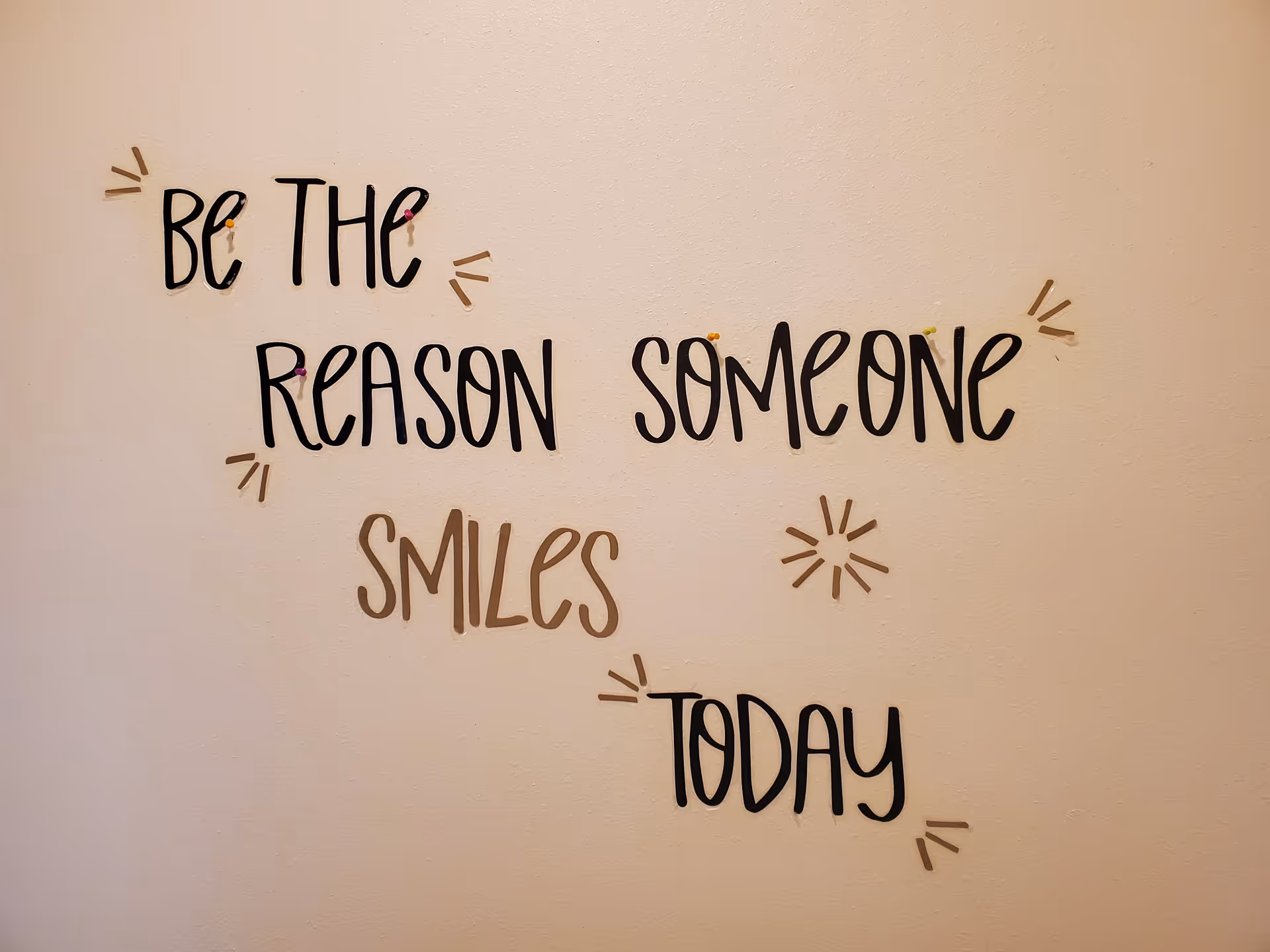 A wall with decorative text that reads 'Be the reason someone smiles today' in black and gold letters with small decorative marks around the words.