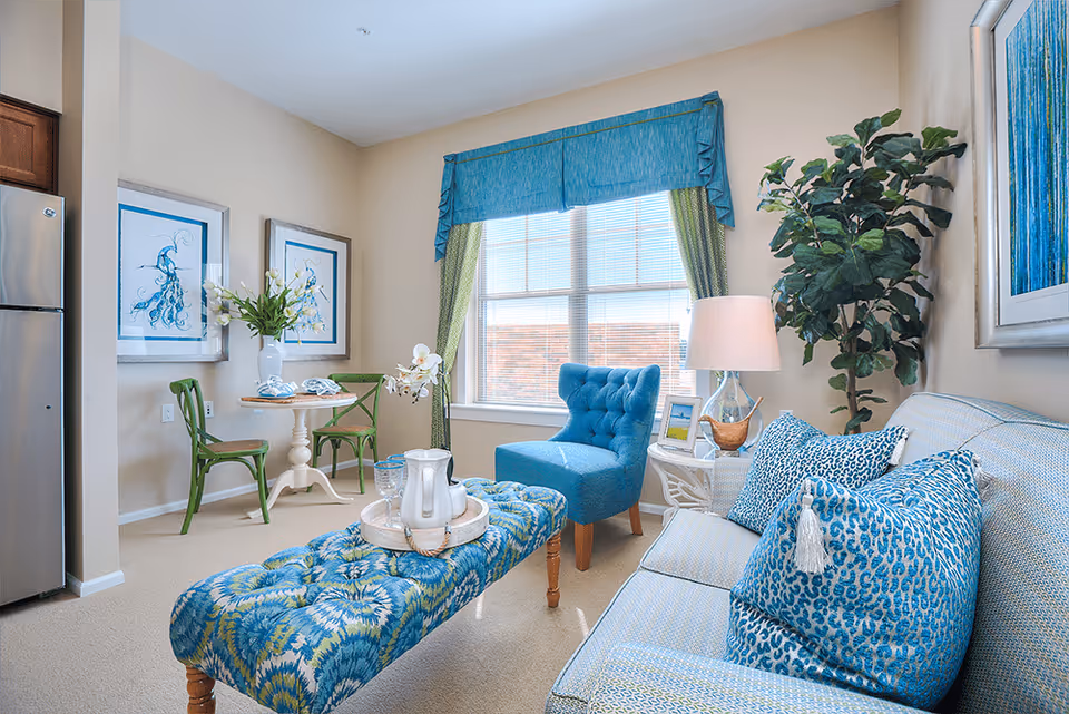 Sunlit living room and dining nook with blue-green upholstered sofa and bench, a tufted accent chair, a small round table with two green chairs, and a large window.