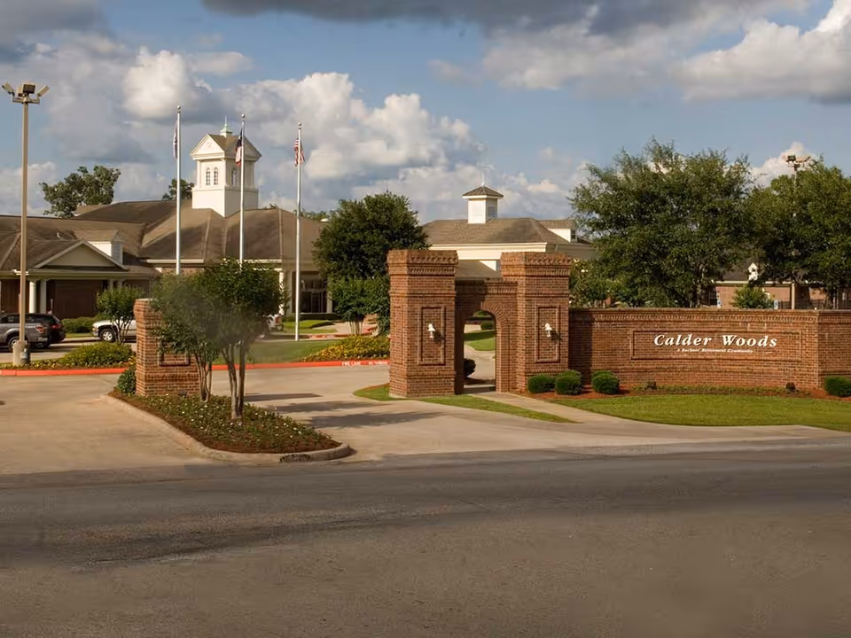 Brick entrance gate and sign for Calder Woods in front of the community building with flagpoles and landscaped grounds.
