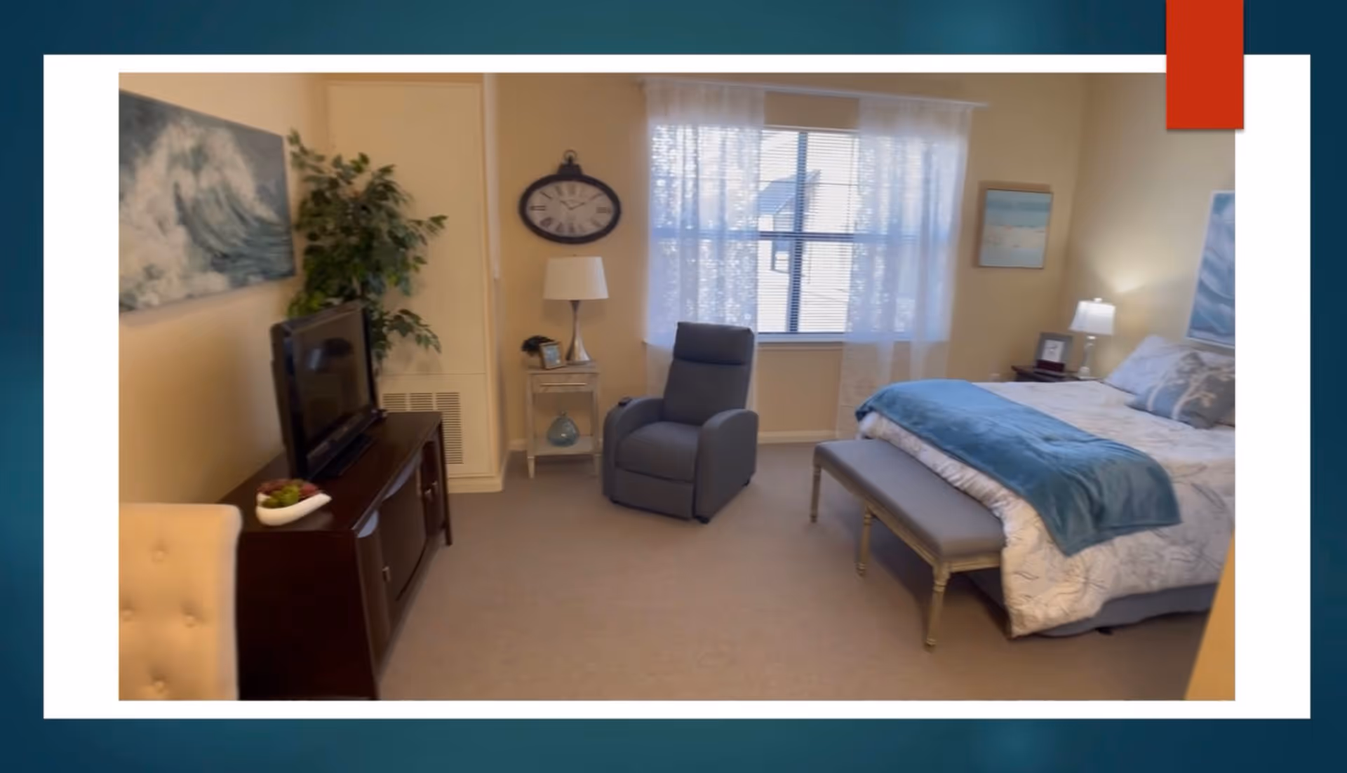 A cozy bedroom with a bed covered in white and blue bedding, a gray bench at the foot of the bed, a gray recliner chair near a window with sheer white curtains, a wooden TV stand with a television, a side table with a lamp and clock, and wall art including a painting of ocean waves and a clock.
