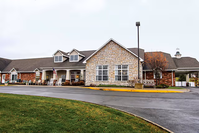 Exterior view of a single-story senior living facility building with a combination of brick and stone facade, multiple windows, a covered porch with rocking chairs, an American flag, and a curved driveway with a grassy area in front under an overcast sky.