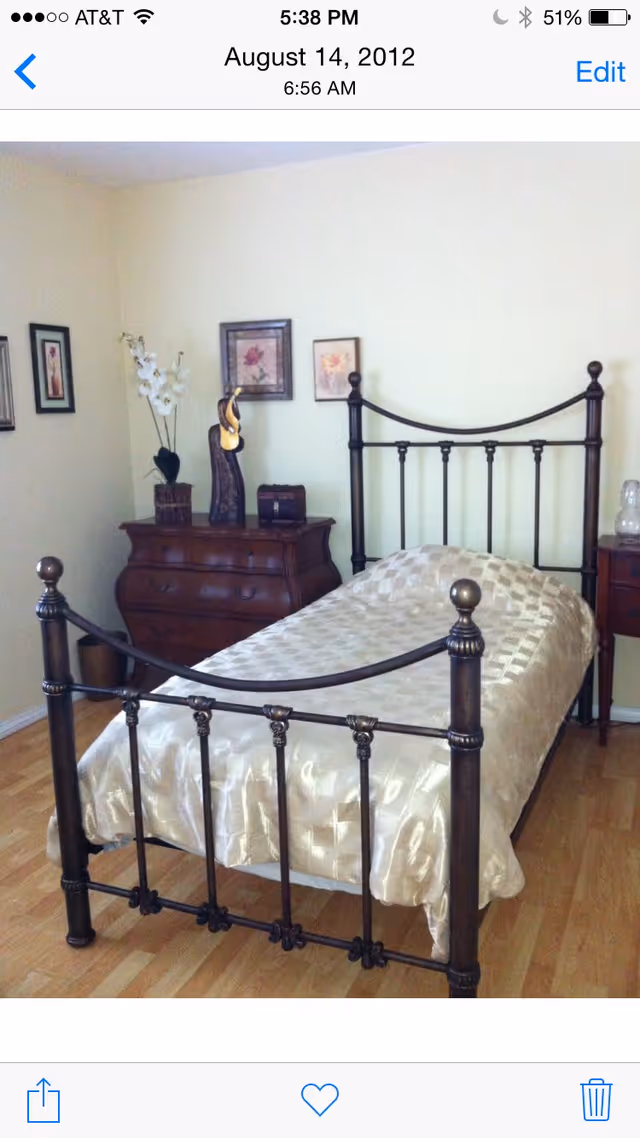 A bedroom with a metal-framed single bed covered with a shiny beige bedspread. Next to the bed is a wooden dresser with decorative items including a vase with white flowers and framed pictures hanging on the wall above it. The floor is wooden and the walls are painted light yellow.
