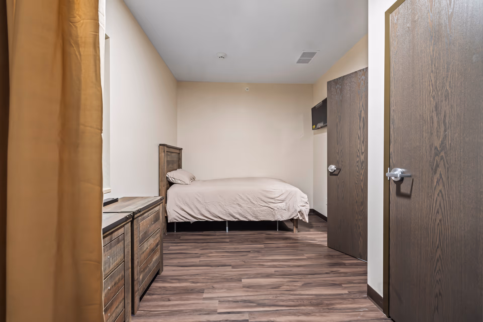 A simple bedroom with a single bed covered in beige bedding against a cream-colored wall. There is a wooden dresser on the left side and two closed wooden doors on the right. The floor is wood laminate, and a small wall-mounted TV is visible near the far door.