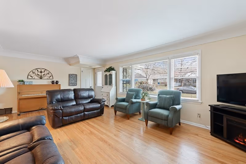 A bright living room with wooden flooring featuring two dark brown leather sofas, two green armchairs, a wooden piano against the wall, a white cabinet, a flat-screen TV on a black stand, and large windows letting in natural light.