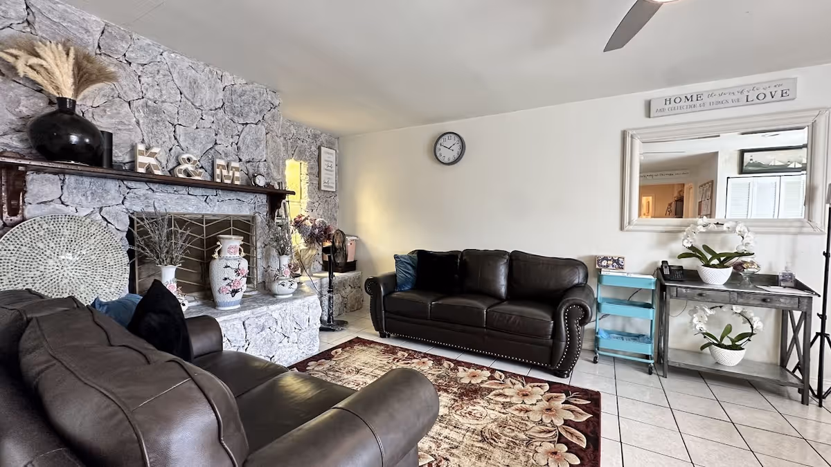 A cozy living room with two dark brown leather sofas facing each other, a floral patterned area rug on white tiled floor, and a stone fireplace decorated with vases and ornamental letters K & M. A wall clock is mounted on a white wall above one sofa, and a large mirror with a decorative sign above it hangs on the adjacent wall. A small table with white orchid plants and a turquoise rolling cart are placed beneath the mirror.