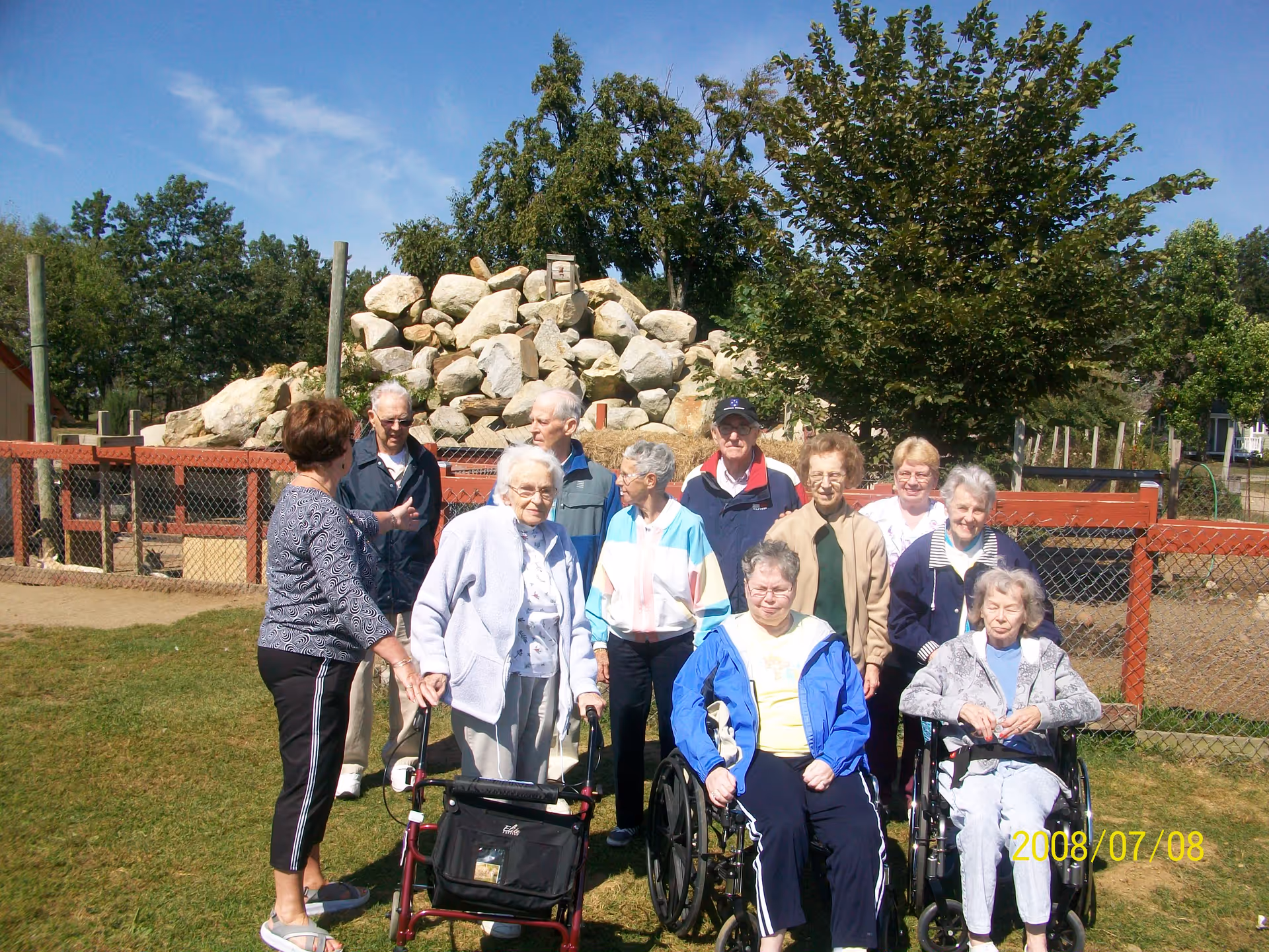 A group of elderly people, some standing and some in wheelchairs, gathered outdoors on a grassy area with a rock pile and trees in the background under a clear blue sky.
