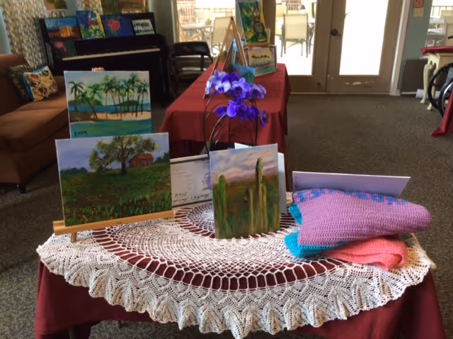 A display table covered with a white crocheted tablecloth and a red cloth underneath, featuring several small paintings on easels, a purple orchid flower, and folded knitted blankets in pink and purple. In the background, there is a brown couch, a piano, chairs, and a door leading outside.