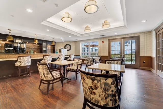 A bright dining room with several tables and floral upholstered chairs arranged neatly. The room features wood flooring, a ceiling with recessed lighting and hanging fixtures, and large windows and glass doors letting in natural light. A counter area with coffee machines and pendant lights is visible in the background.