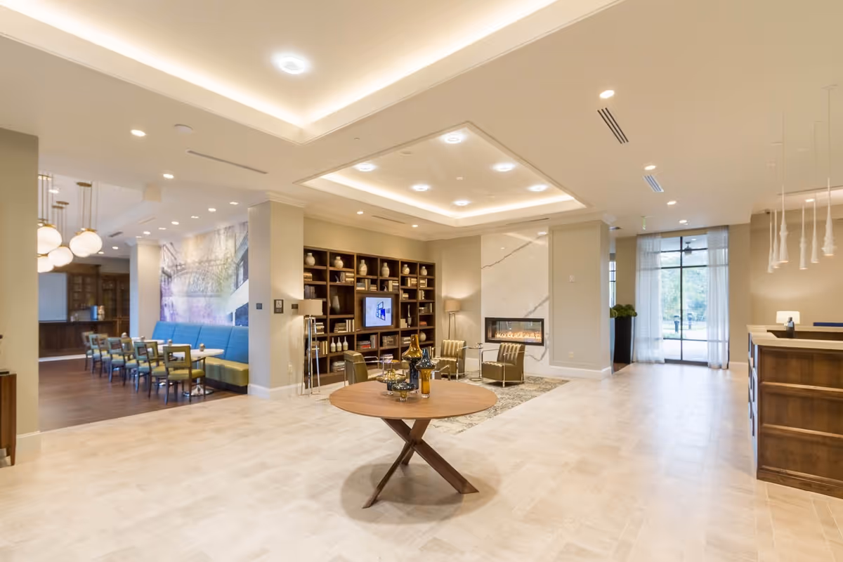 Bright modern lobby/common area with seating, a round table, fireplace, bookshelf and a dining area visible to the left.