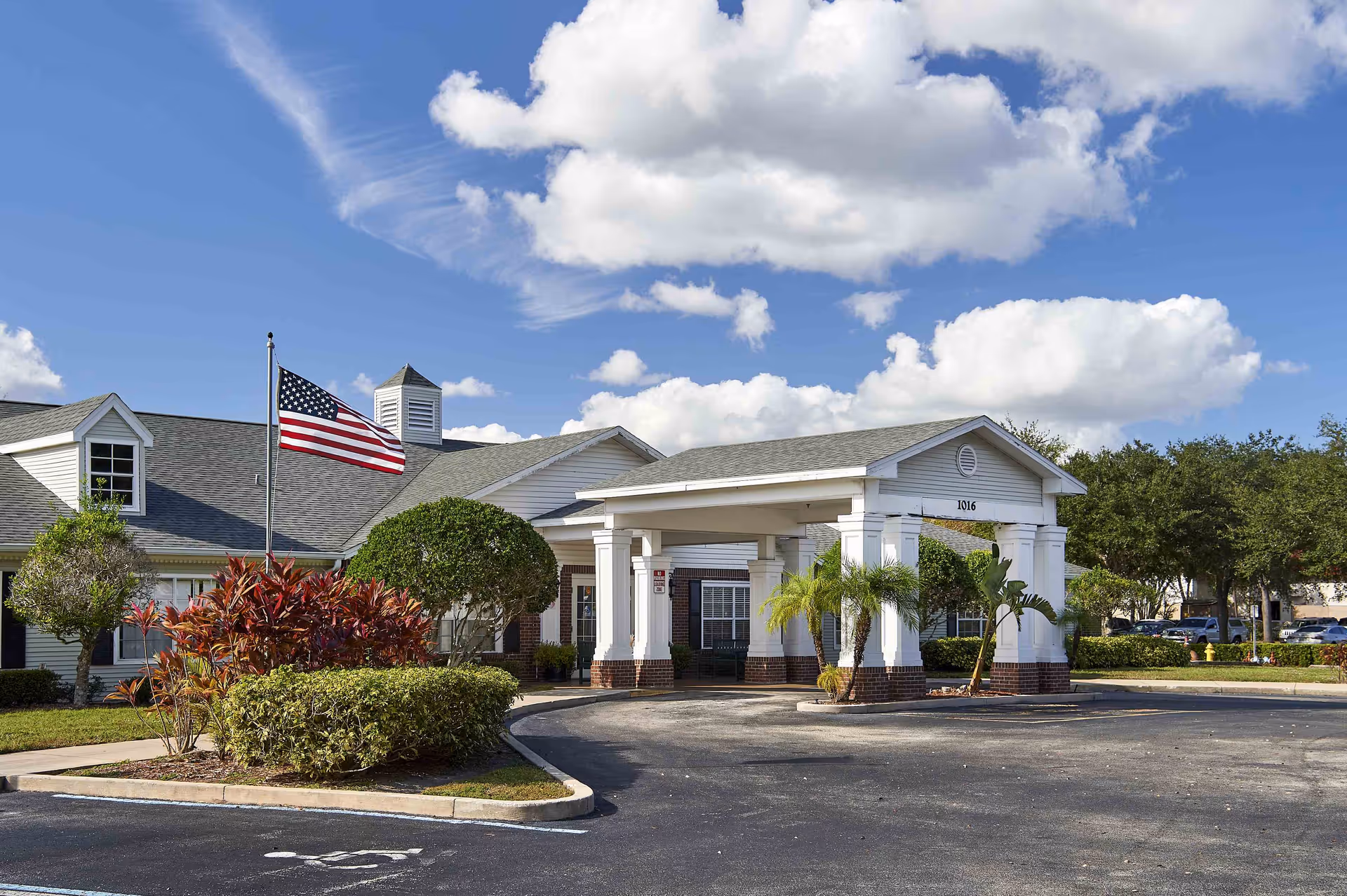 Exterior view of Palmetto Landing facility showing a single-story building with a covered entrance, an American flag on a flagpole, well-maintained landscaping with bushes and small trees, and a partly cloudy blue sky.