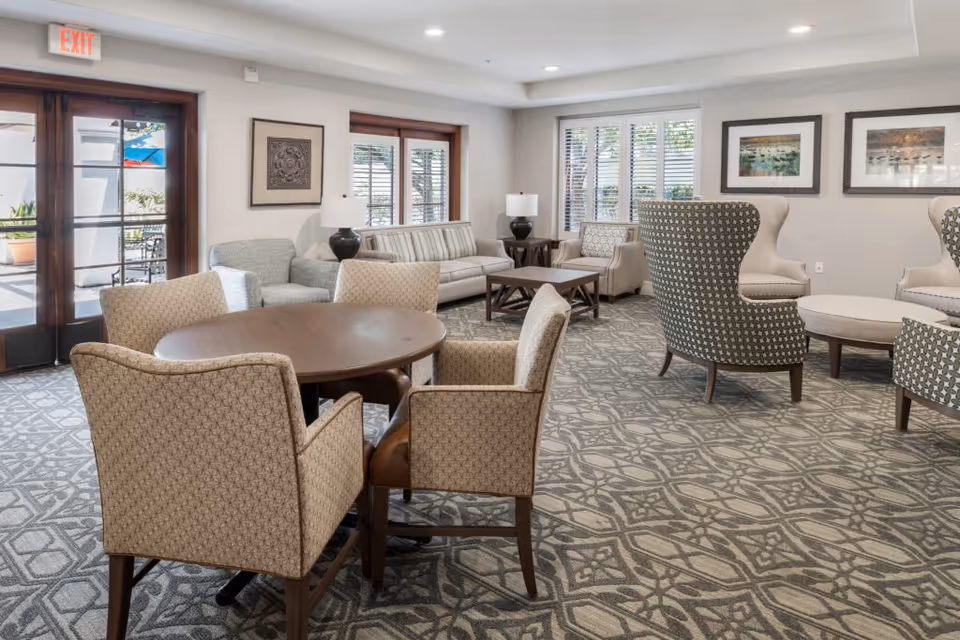 A bright and spacious living room area with patterned carpet, a round wooden table surrounded by four upholstered chairs, several armchairs, a sofa, side tables with lamps, framed artwork on the walls, and large windows with white blinds letting in natural light. A glass door with an exit sign leads outside to a patio area.
