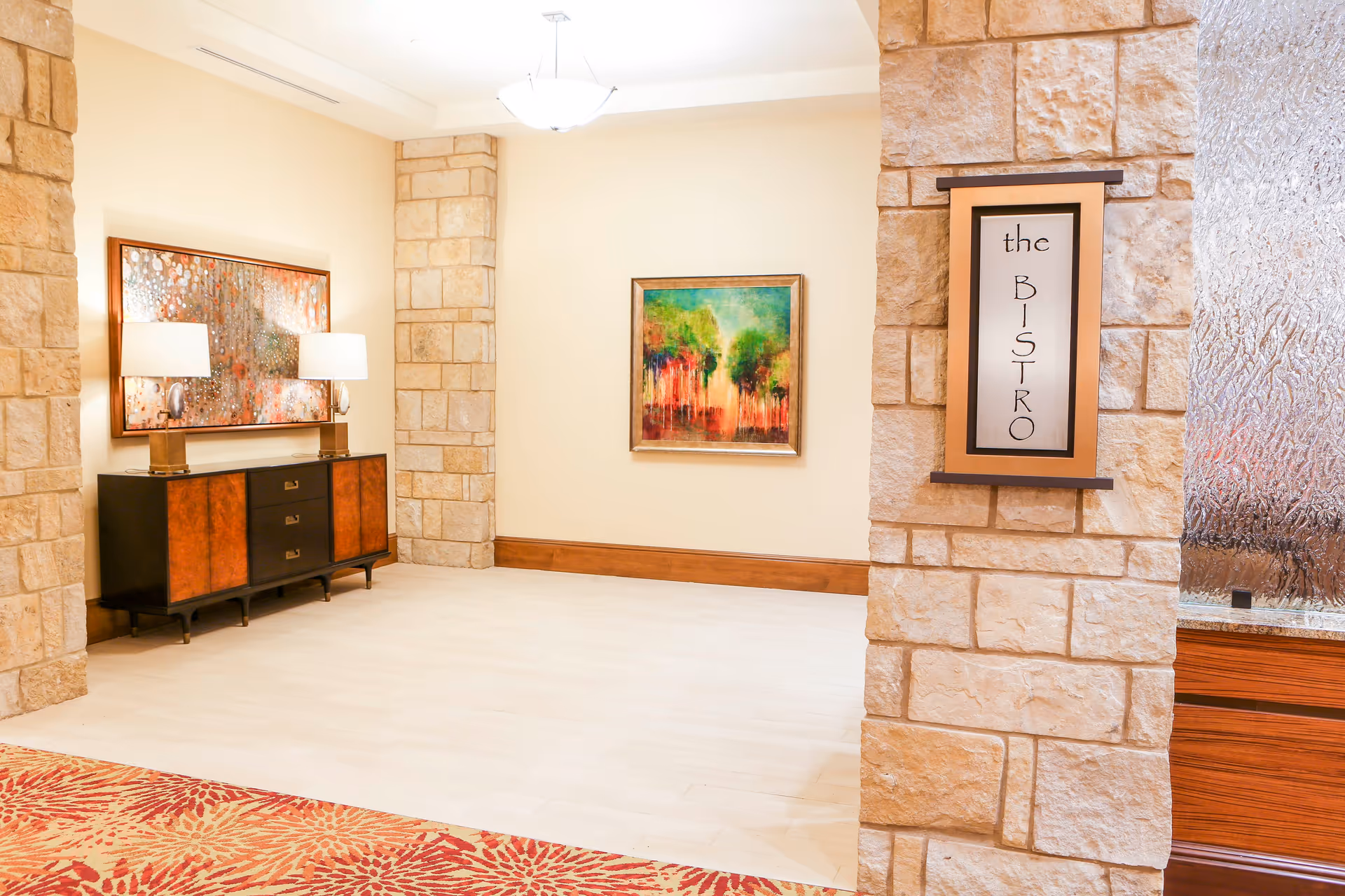 Well-lit interior lobby with stone pillars, a console table and lamps, colorful artwork, and a sign reading "the BISTRO".