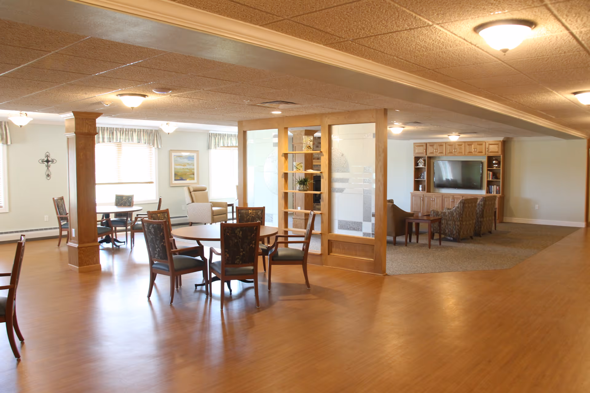 A spacious senior living common area with wooden floors and ceiling tiles. The room features several round tables with chairs, a cozy seating area with armchairs facing a large flat-screen TV mounted on a wooden entertainment center, and large windows letting in natural light. A wooden partition with glass panels and shelves separates the seating area from the dining area.