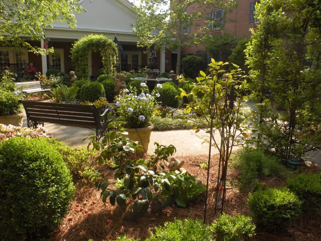 A lush garden area with various green shrubs, flowering plants, and small trees. There are benches for seating and decorative statues scattered throughout. The garden is adjacent to a building with a covered porch and brick walls, bathed in sunlight.