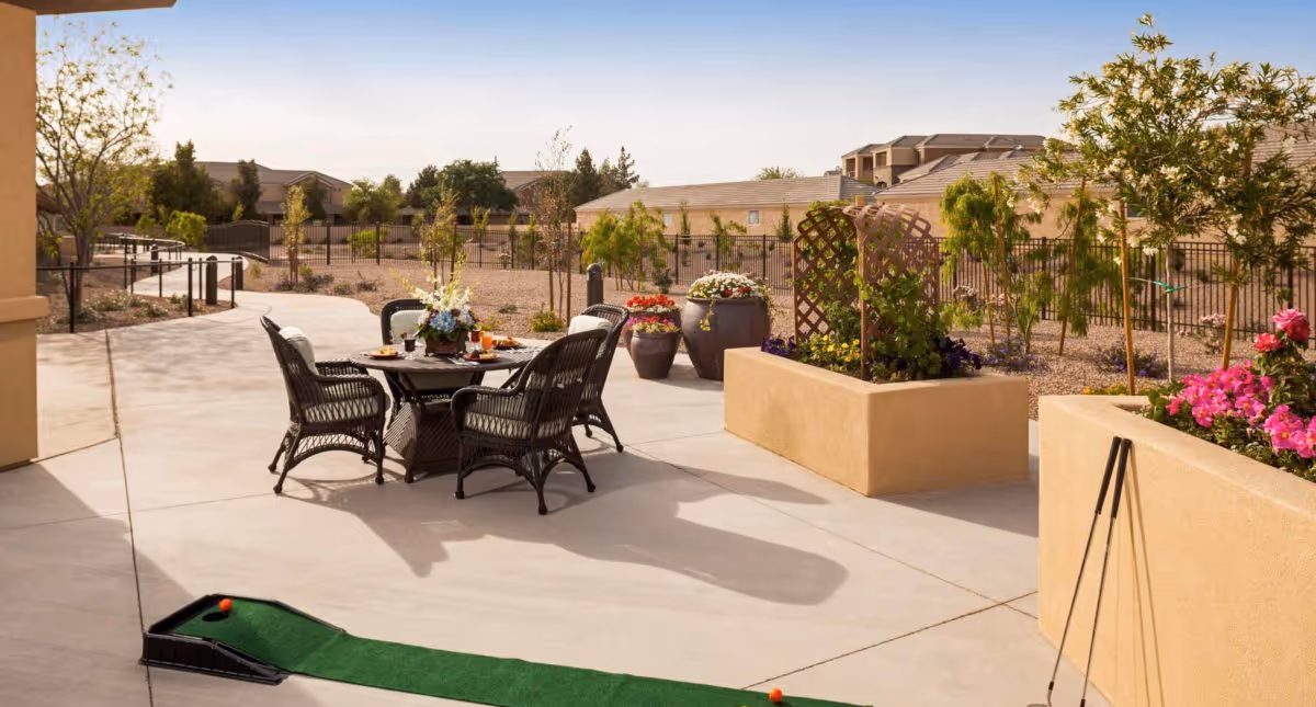 Outdoor patio area with a round table and four wicker chairs, set with plates and a flower centerpiece. There are potted plants and raised garden beds with flowers and greenery. A small putting green with golf clubs and balls is visible in the foreground. The background shows a fenced garden area and residential buildings under a clear sky.