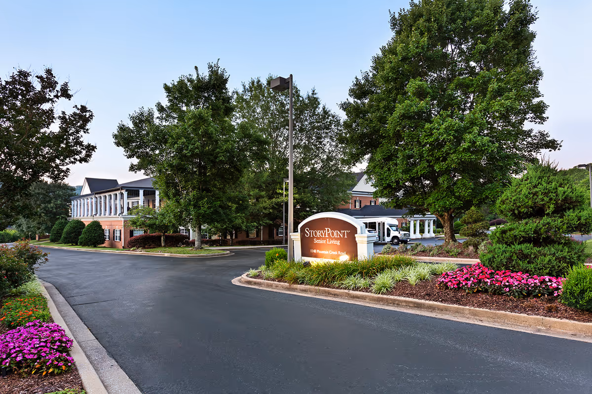 Entrance driveway to StoryPoint Senior Living facility with a large sign displaying the name and address, surrounded by landscaped greenery and flowers, with the building partially visible behind trees under a clear sky.