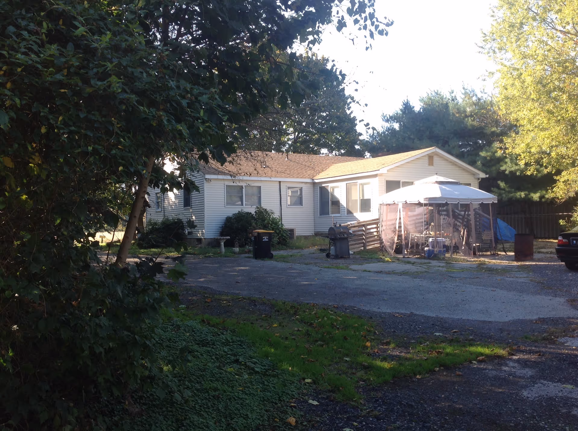 Exterior view of a single-story white house with a brown roof, surrounded by trees and greenery. There is a paved area in front with a barbecue grill, a canopy tent with outdoor furniture underneath, and a parked car partially visible on the right side.