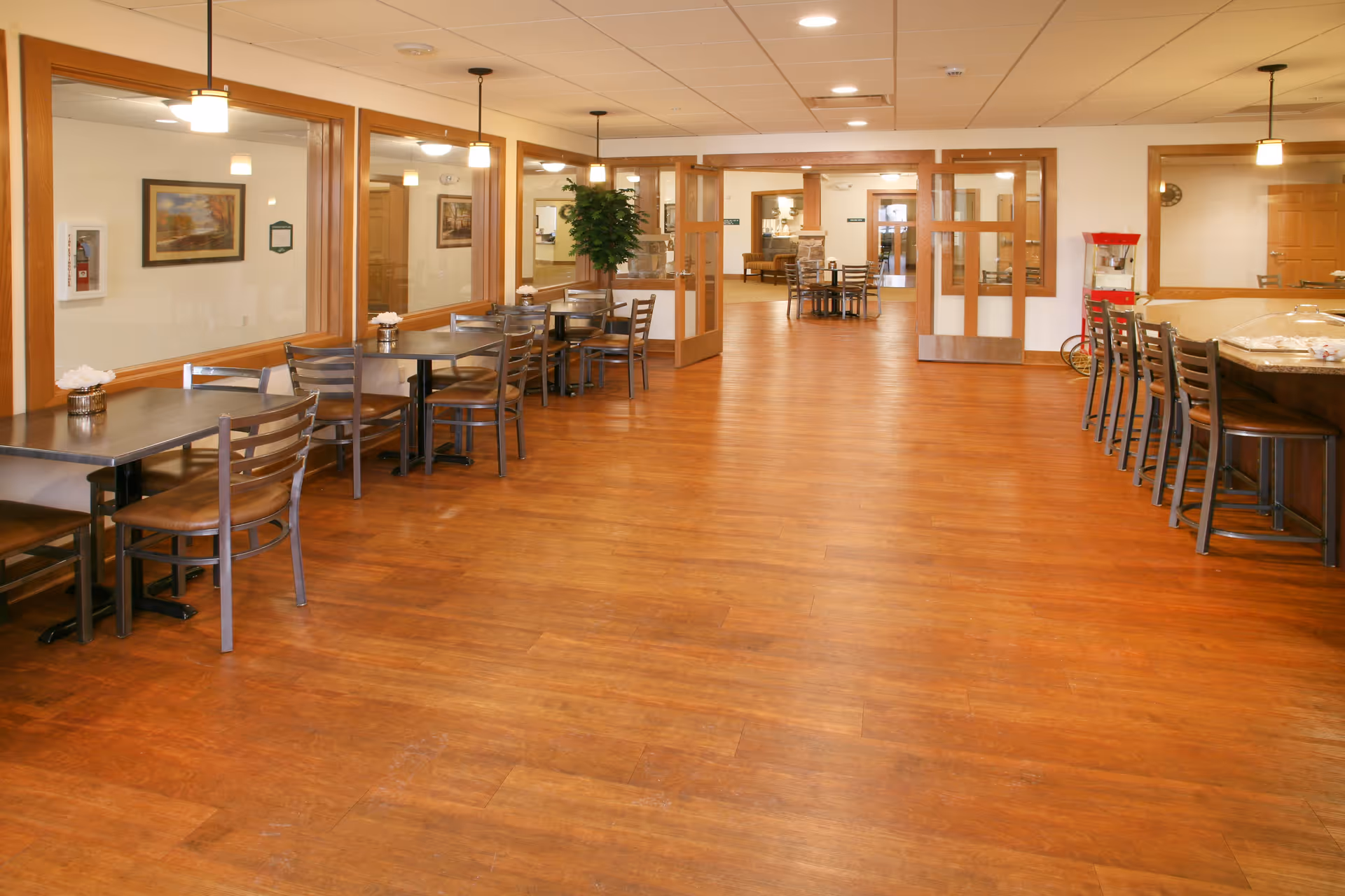 A spacious dining area with wooden floors and multiple tables and chairs arranged along the walls and in the center. The room features large windows with wooden frames, pendant lights hanging from the ceiling, and a popcorn machine on the right side. The area is well-lit with ceiling lights and has a warm, inviting atmosphere.