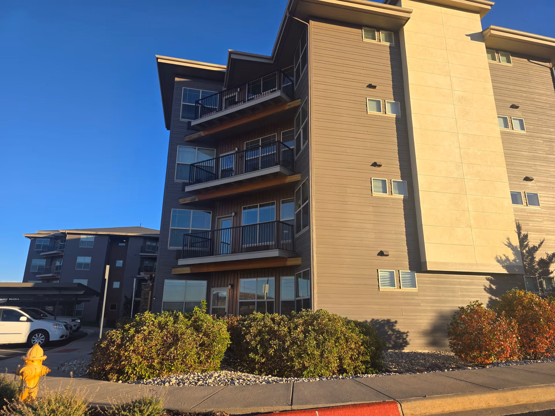 Exterior view of a multi-story residential building with balconies, surrounded by bushes and a sidewalk. A yellow fire hydrant is visible in the foreground, and several parked cars are seen to the left under a carport. The sky is clear and blue.